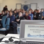 Laptop-Monitor mit PowerPoint-Folie im Vordergrund, Teilnehmer im Hintergrund