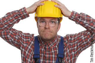 Bauarbeiter mit gelbem Helm, Karohemd und Brille fasst sich vor Verzweiflung an den Kopf