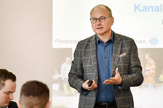 Mann mit Brille, blauem Hemd und kariertem Sakko hält Vortrag